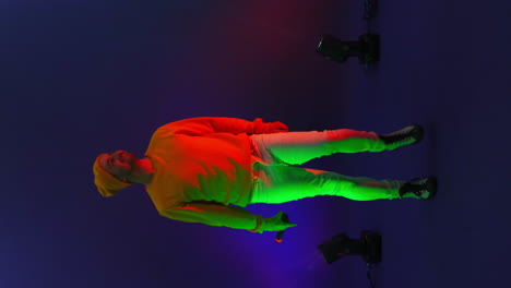 Vertical-video.-A-man-in-a-yellow-jacket-dancing-on-a-blue-background.-The-singer-in-the-Studio-in-the-neon-light-dancing-and-singing.-Changing-the-background-color.-Strobe-lights-and-funny-dancer